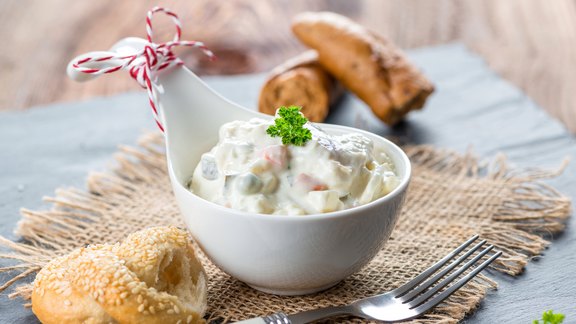 Hausgemachter Heringssalat in einer weißen Porzellanschüssel, garniert mit Petersilie. Serviert auf rustikalem Jutetuch mit frischem Sesamgebäck und einer Gabel. Die Schüssel ist mit einer dekorativen rot-weiß gestreiften Kordel verziert.  